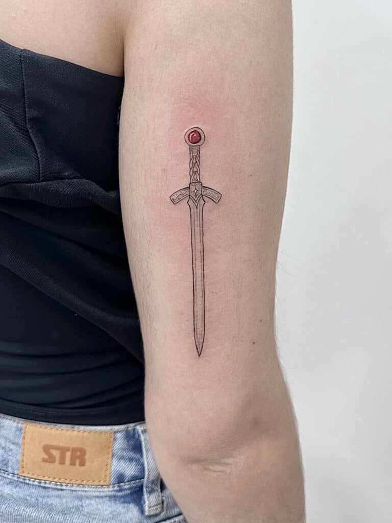 Tatuaje de línea fina de espada con detalle de piedra en rojo en el brazo