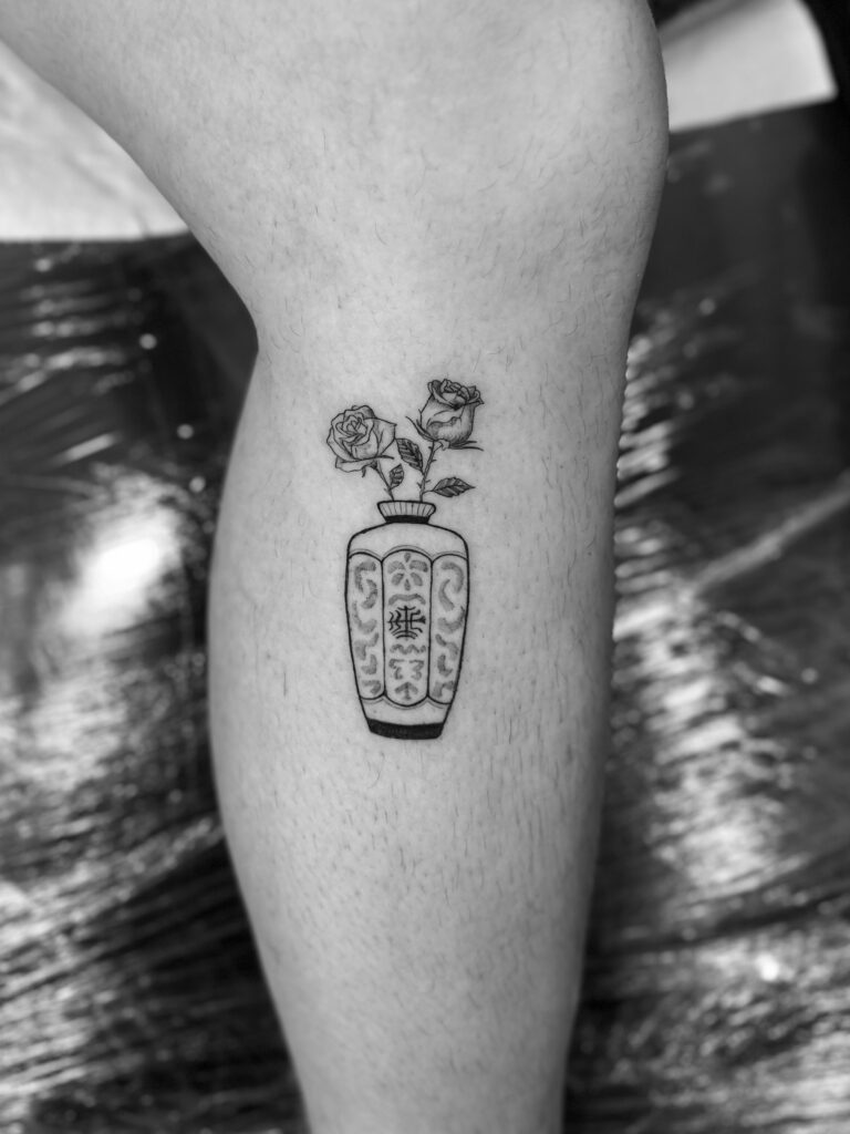 Tatuaje en línea fina de un jarrón de inspiración asiática con una rosa en la pierna