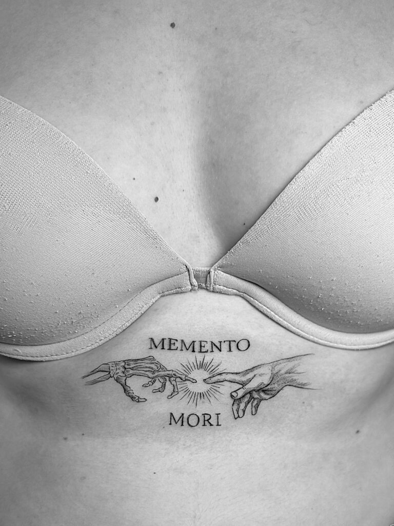 Tatuaje en línea fina de frase "Memento Mori" con dibujo de Miguel Ángel en el pecho